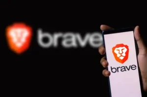 Promotional image showing a hand holding a smartphone with the Brave browser logo—an orange lion head above the word “brave”—with a blurred larger logo in the background.